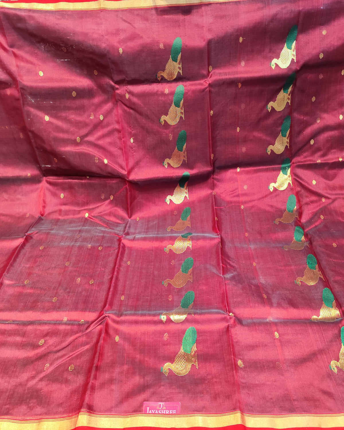 A deep red silk saree with gold zari embroidery, featuring green and gold floral motifs and a red and gold border, displayed flat on a table with fabric stacks in the background.