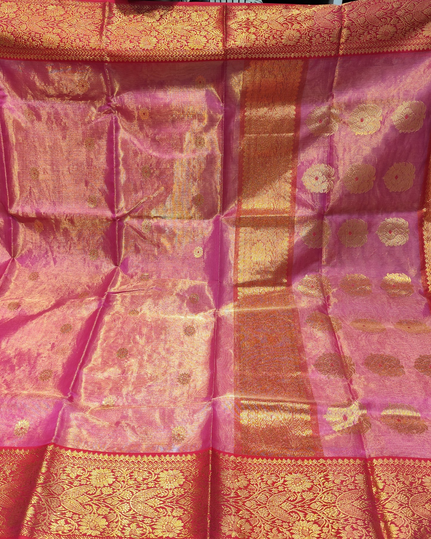 Chanderi Tissue Silk Pink silk saree is held up to display its intricate woven patterns and borders. The fabric features a rich magenta-pink base with metallic gold zari work, including a wide decorative border and small circular motifs.