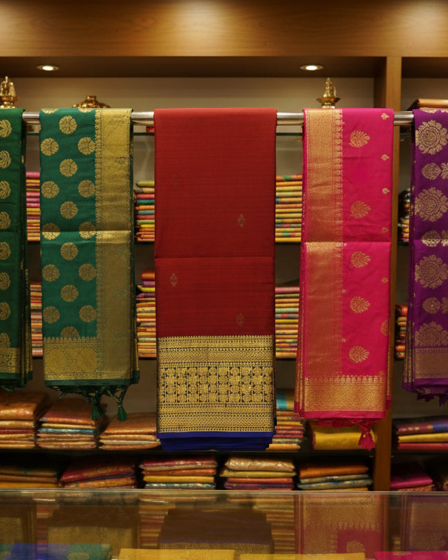 A folded red silk saree with a wide gold and black zari border and a blue underlayer visible at the bottom, displayed on a light surface.