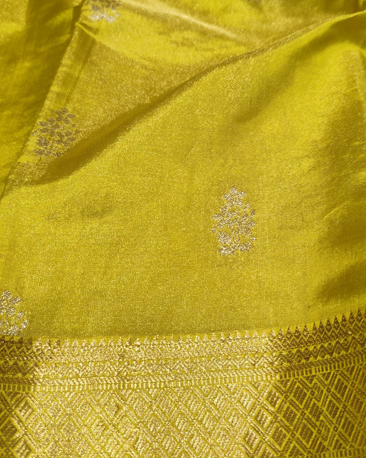 A close-up of a bright yellow-green silk fabric, likely a saree, featuring a wide gold zari brocade border and small metallic floral motifs woven throughout.