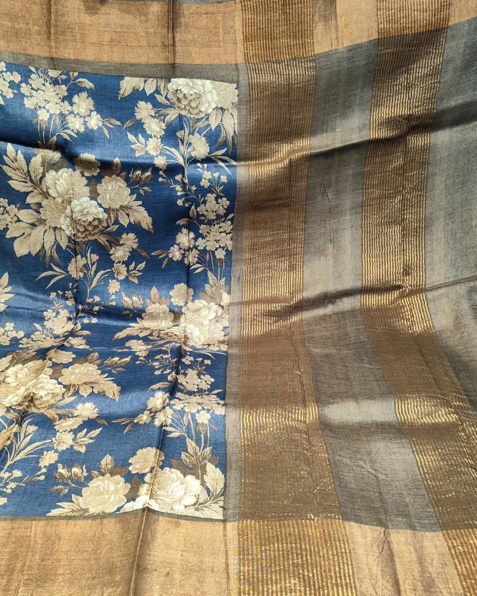 A close-up of a blue and beige floral Tussar silk saree showing the detailed floral pattern on the body and the striped gold and gray border.