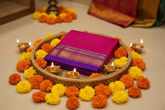 A folded magenta pink fabric garment, likely a sari or dress, featuring a wide purple border with intricate gold and orange woven patterns, laid flat on a white surface.