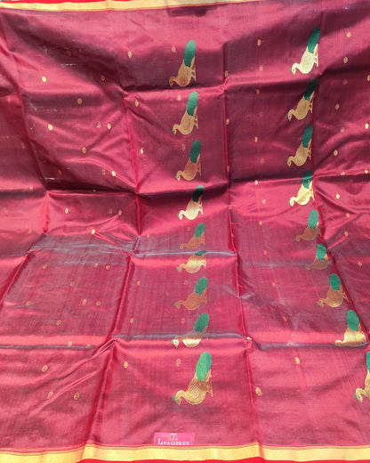 A deep red silk saree with gold zari embroidery, featuring green and gold floral motifs and a red and gold border, displayed flat on a table with fabric stacks in the background.