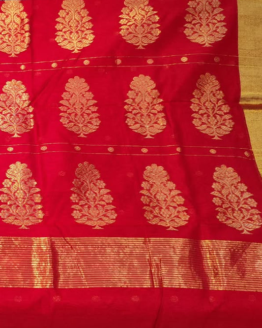 A person holds up a red and gold woven fabric, likely a saree or scarf, featuring a rich red body with gold zari motifs and borders, set against a background of folded fabrics.