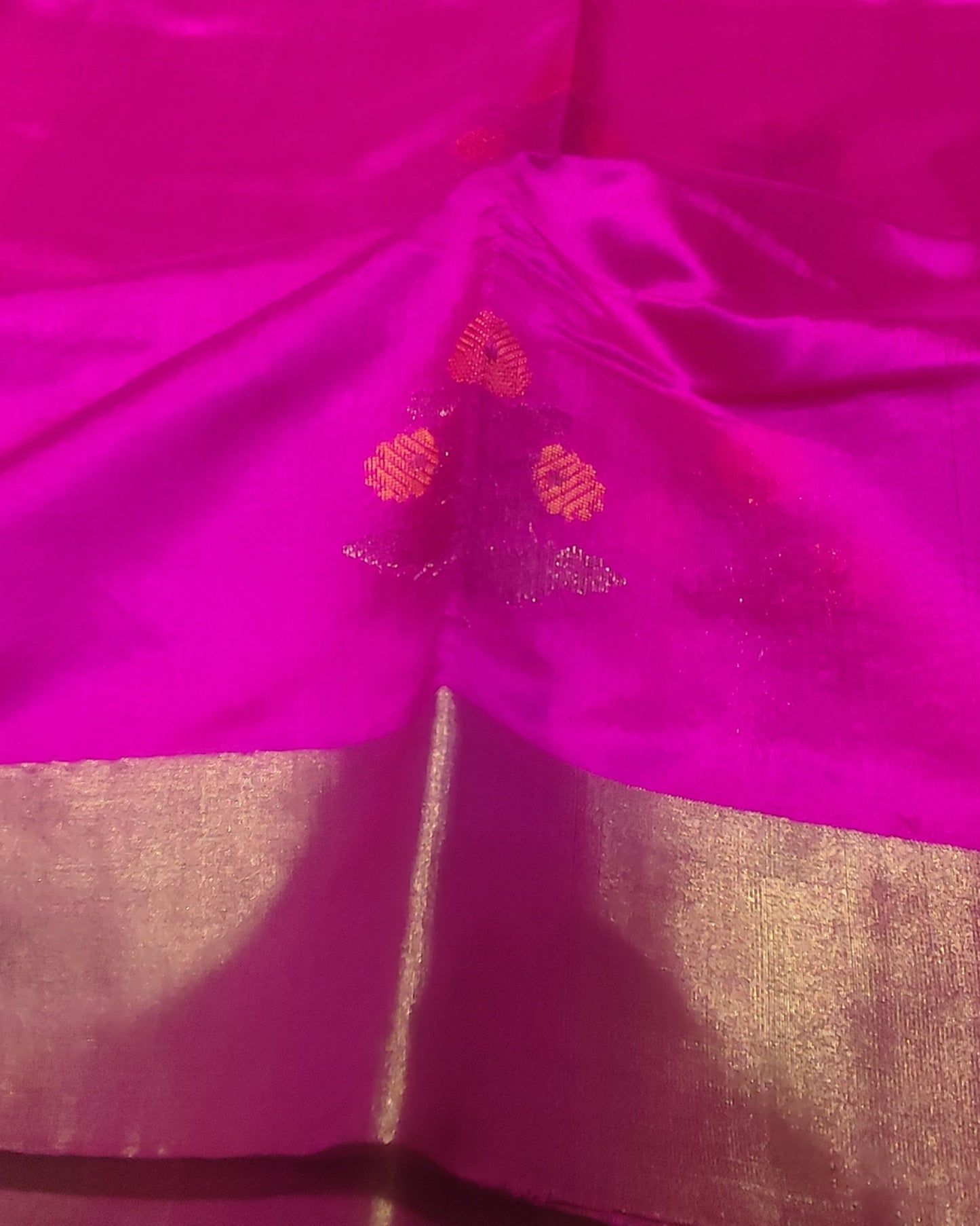 A close-up of a bright magenta pink fabric, likely silk or satin, featuring a wide metallic gold border and small gold woven floral motifs.