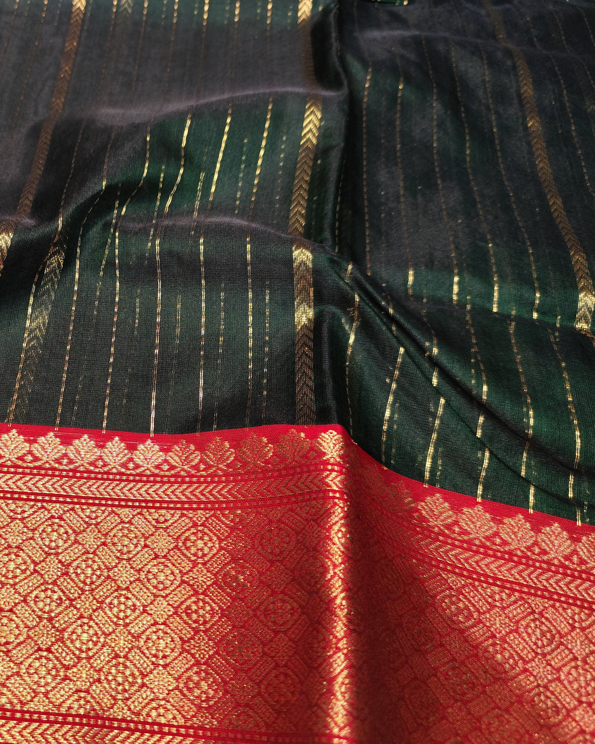 A close-up of a dark green and black silk saree fabric with vertical gold stripes and a wide red border featuring intricate gold zari woven patterns.