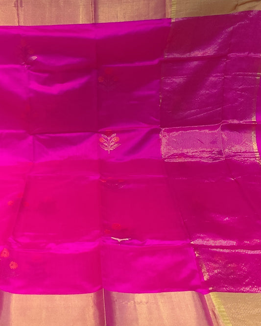 A bright fuchsia pink silk saree with a wide gold zari border, displayed flat against a background of other folded fabrics.