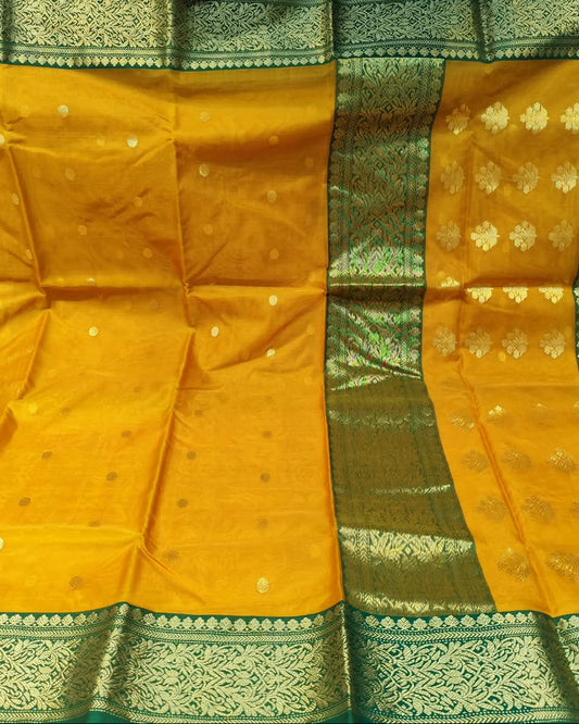 A yellow and green silk saree with gold zari brocade work, displayed flat. The fabric features a solid yellow body with small gold motifs, a wide green border with gold detailing, and a vertical panel of gold floral motifs on the green background.