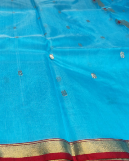 A bright turquoise blue fabric, likely a saree or scarf, featuring small metallic silver floral motifs scattered across the body and a decorative border at the bottom with red, gold, and blue stripes.