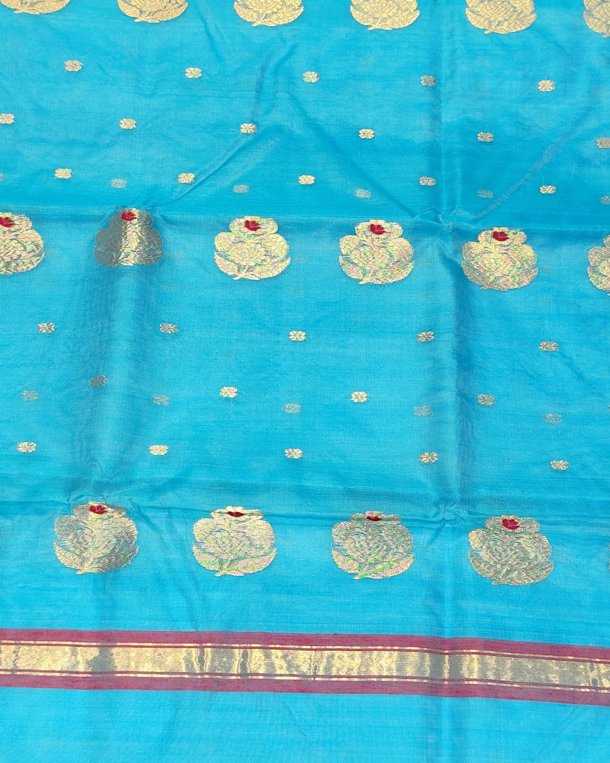 A bright blue saree fabric with metallic gold and silver zari motifs and borders, held up by a person in a shop with stacks of colorful textiles in the background.