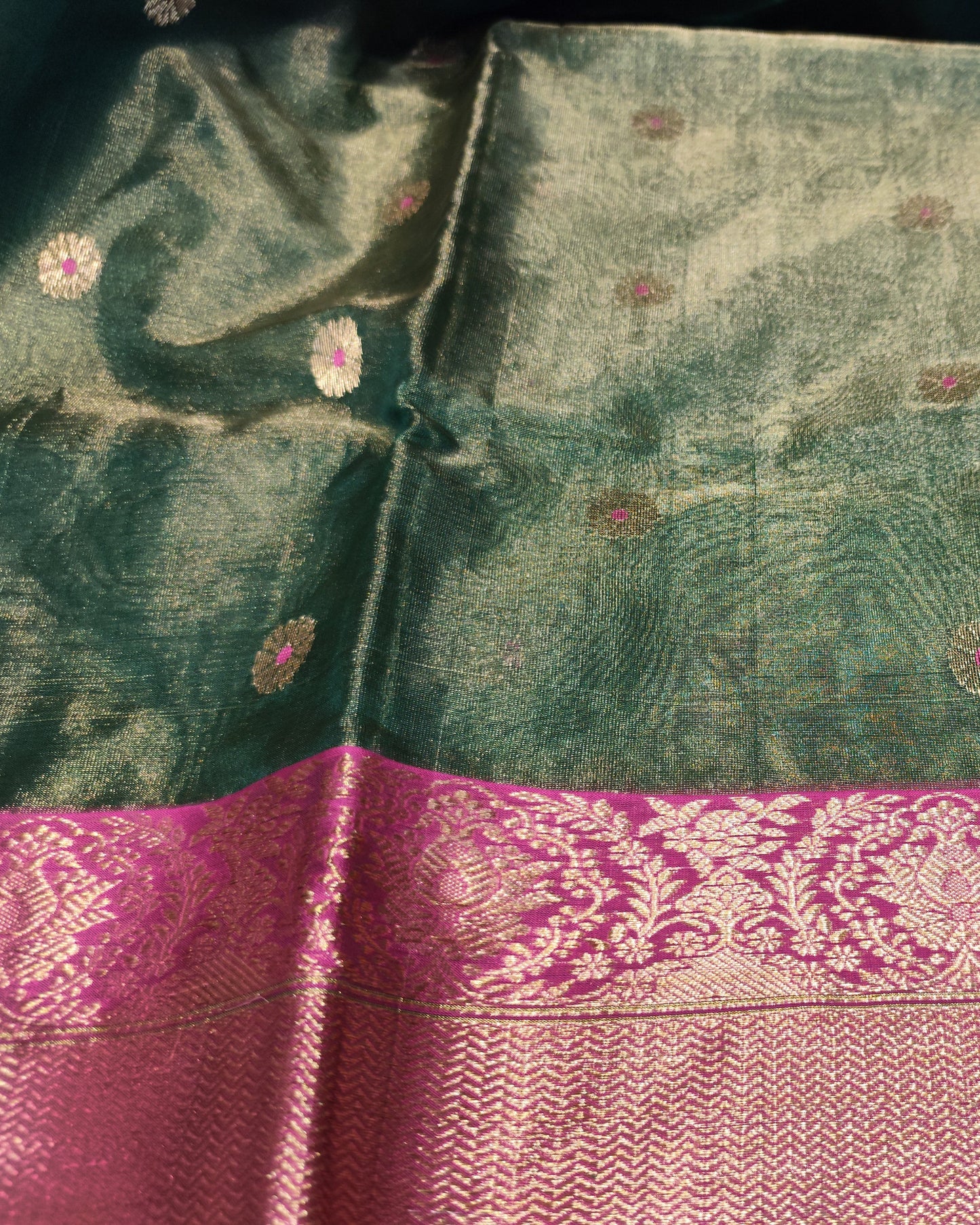 A dark green silk saree with a wide magenta-pink border featuring intricate gold zari woven patterns.