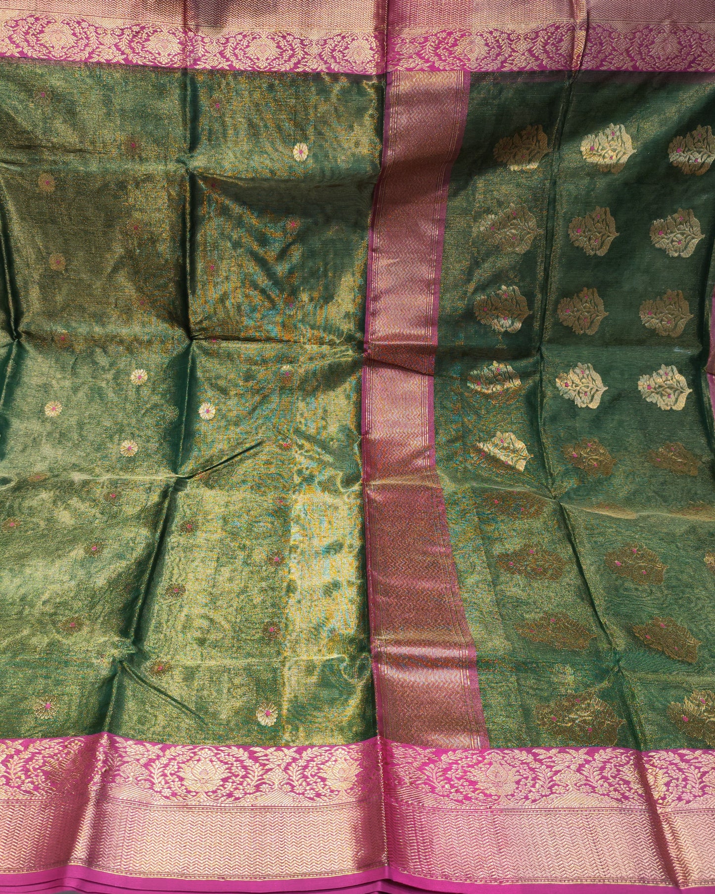 A green silk saree with gold zari floral motifs and a wide pink border, displayed flat against a background of folded fabrics.