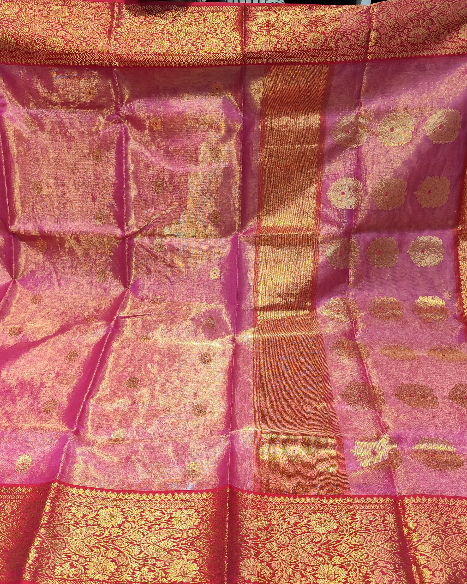 Chanderi Tissue Silk Pink silk saree is held up to display its intricate woven patterns and borders. The fabric features a rich magenta-pink base with metallic gold zari work, including a wide decorative border and small circular motifs.