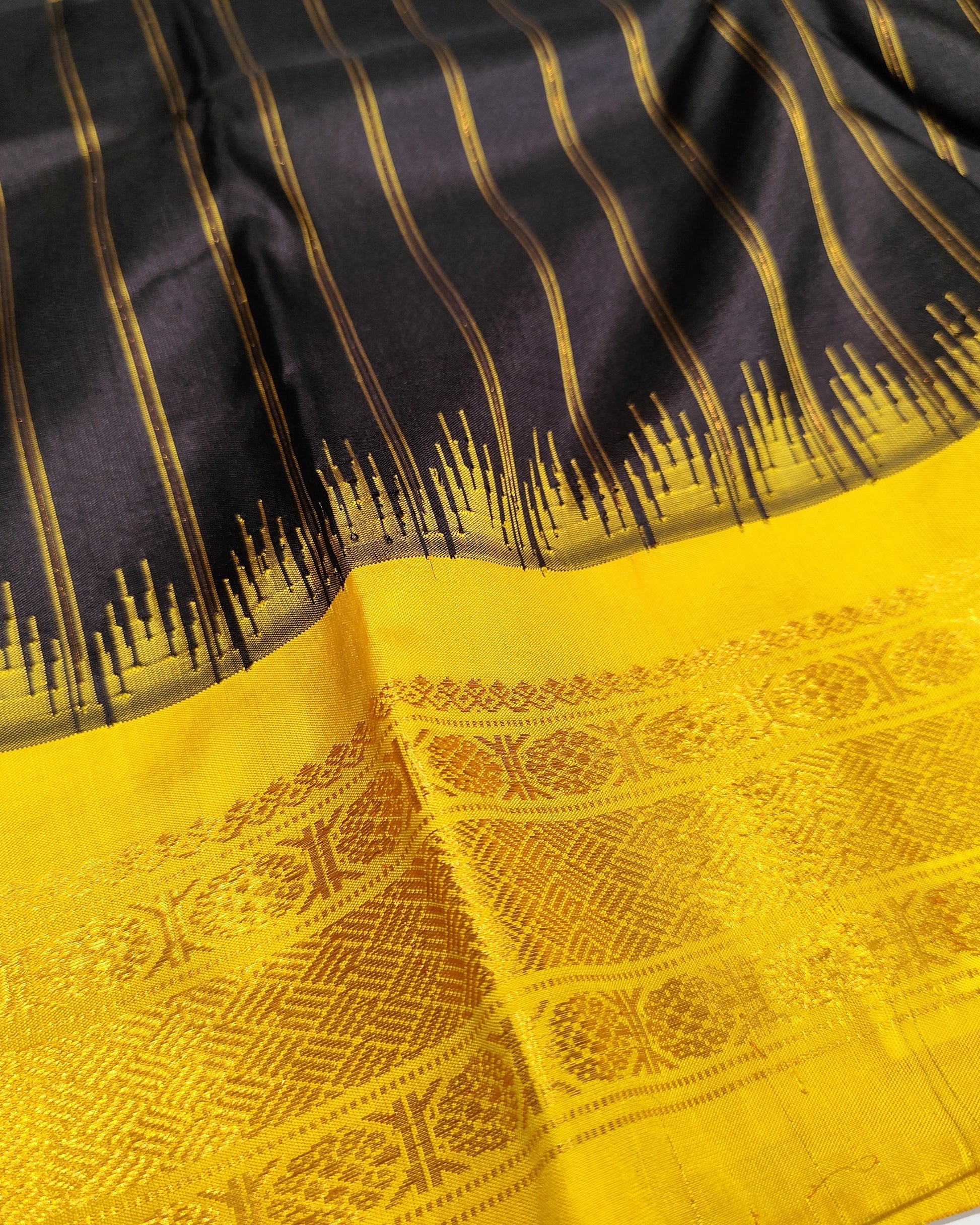 A close-up of a black and yellow woven saree, featuring vertical gold stripes on a black body, a jagged gold border, and a wide yellow pallu with intricate gold woven patterns.