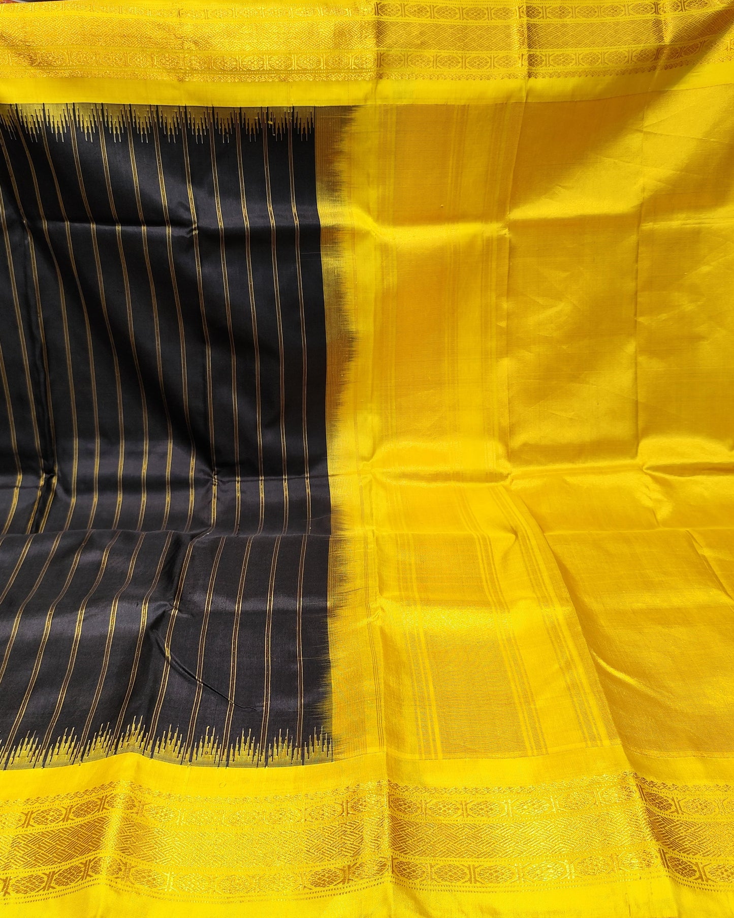 A yellow and black silk saree is held up by hands in a fabric store. The left side of the saree has a black base with fine yellow stripes, while the right side and border are solid yellow with a decorative zari border.