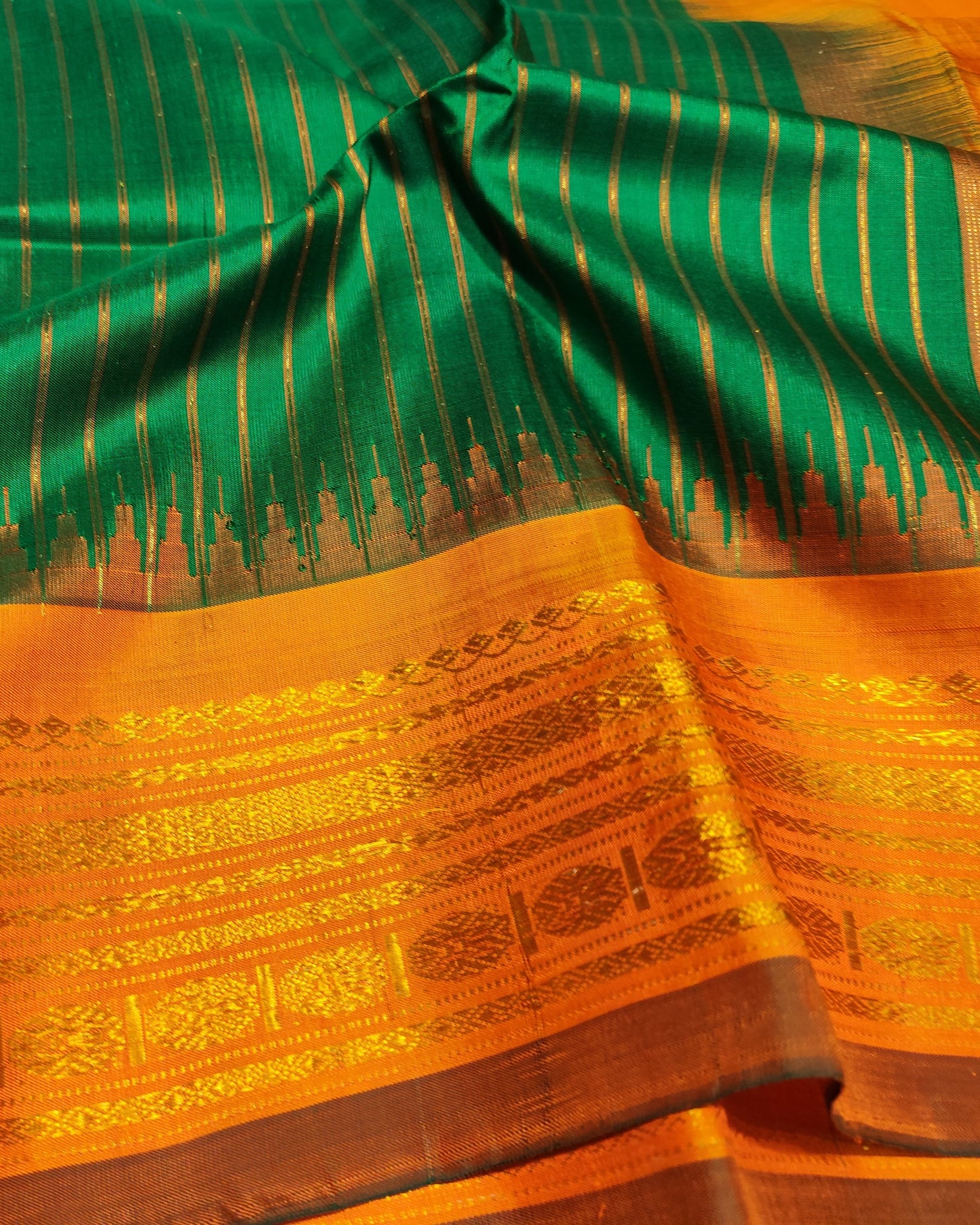 A close-up of a richly patterned Gadwal sari with a vibrant green body featuring vertical metallic gold stripes, a wide golden-orange border with intricate woven motifs, and a dark brown zari edge.