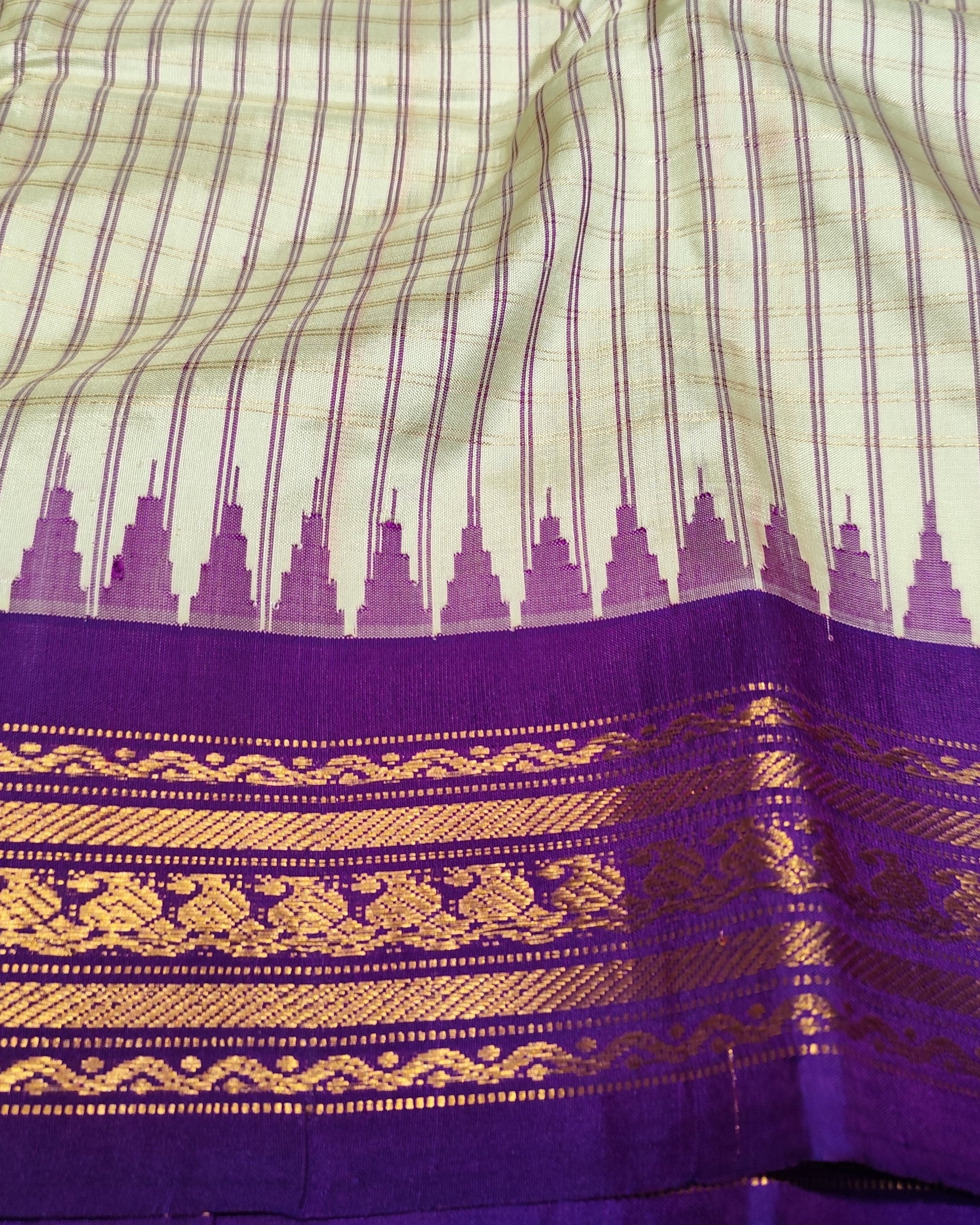 A close-up of a traditional Indian silk saree fabric, showing a cream-colored body with thin purple stripes, a row of purple motifs, and a wide purple border with intricate gold zari weaving.