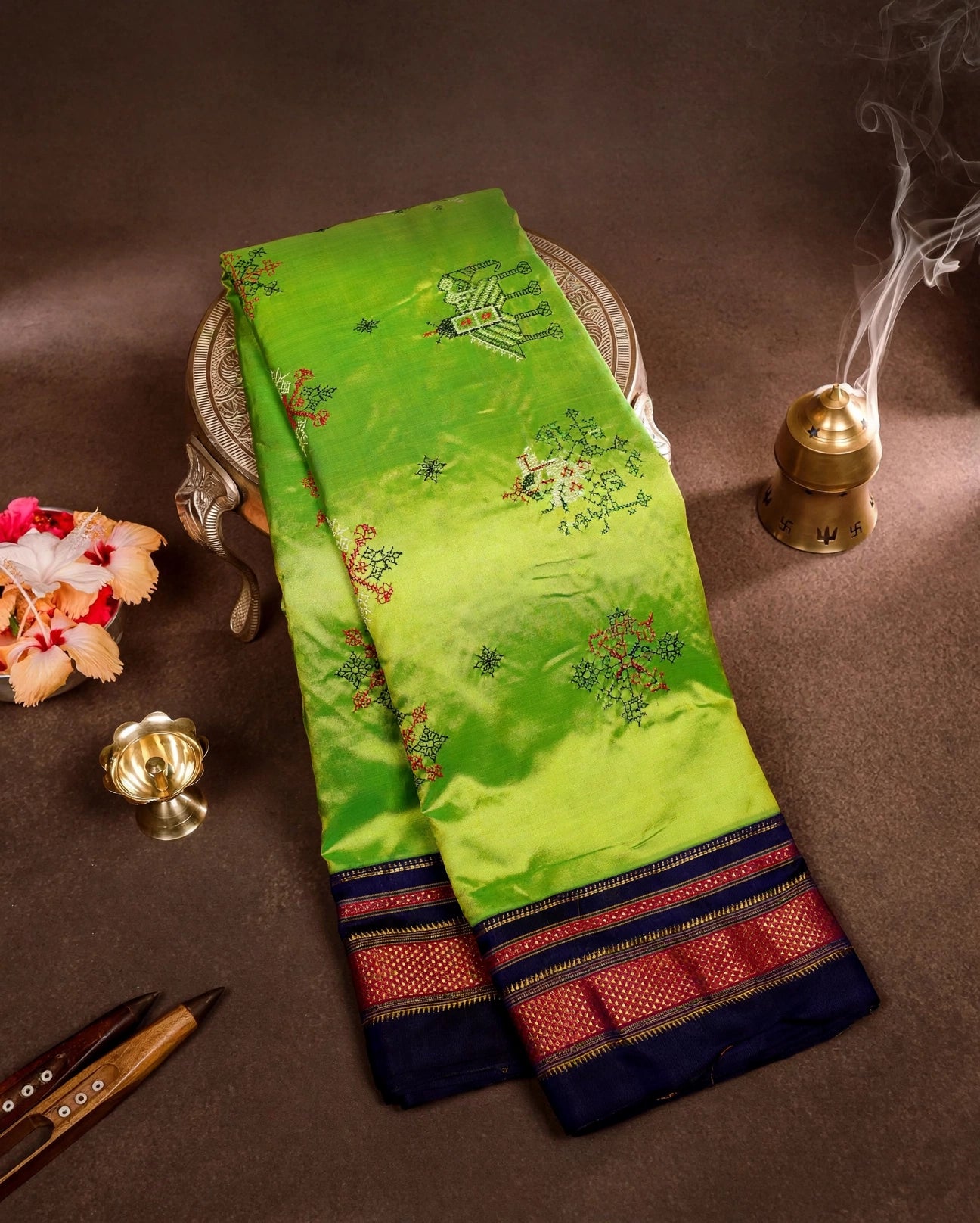 A folded green Ilkal silk saree with floral embroidery and a navy blue border with red and gold stripes, displayed on a brown surface with decorative props.