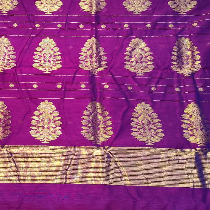 A person holds up a purple silk saree with gold zari motifs and borders in a fabric store.