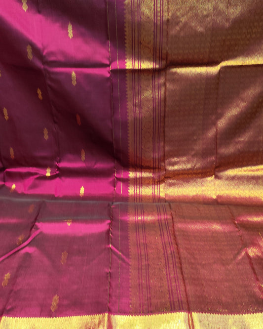 A close-up of a draped silk Kanjeevaram saree fabric, showing its rich magenta-pink body with a wide golden zari border and small gold motifs.