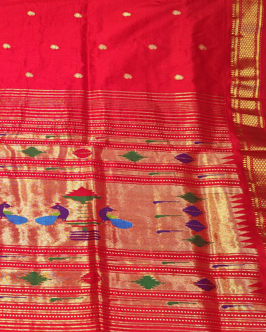 A person holds up a red and gold woven fabric, likely a saree or dupatta, featuring intricate gold zari patterns, small gold dots, and colorful woven accents in blue, green, and purple. The background shows a shop interior with shelves of folded textiles.