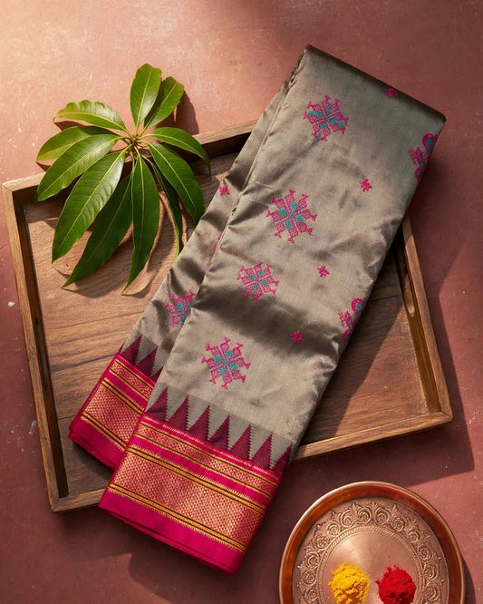 A folded silk gray saree with pink and blue geometric motifs and a pink and gold zari border, displayed on a wooden tray alongside green leaves and a decorative plate with yellow and red powder.