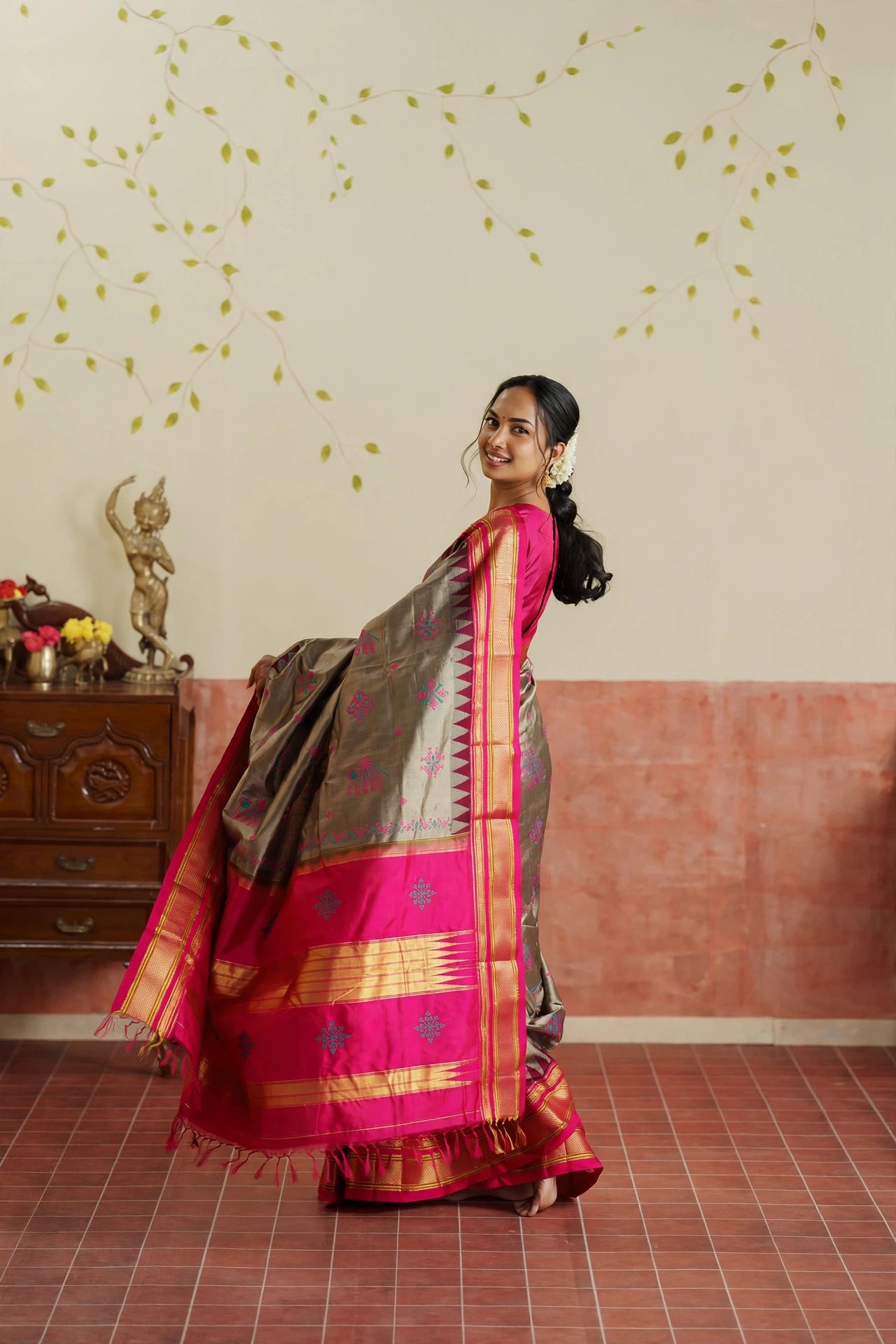 A woman modeling a traditional Indian Ilkal sik grey saree in a decorated indoor room. The saree has a grey body with a vibrant pink border and gold zari detailing, paired with a solid pink blouse.