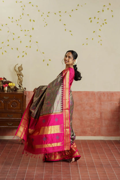 A woman modeling a traditional Indian Ilkal sik grey saree in a decorated indoor room. The saree has a grey body with a vibrant pink border and gold zari detailing, paired with a solid pink blouse.