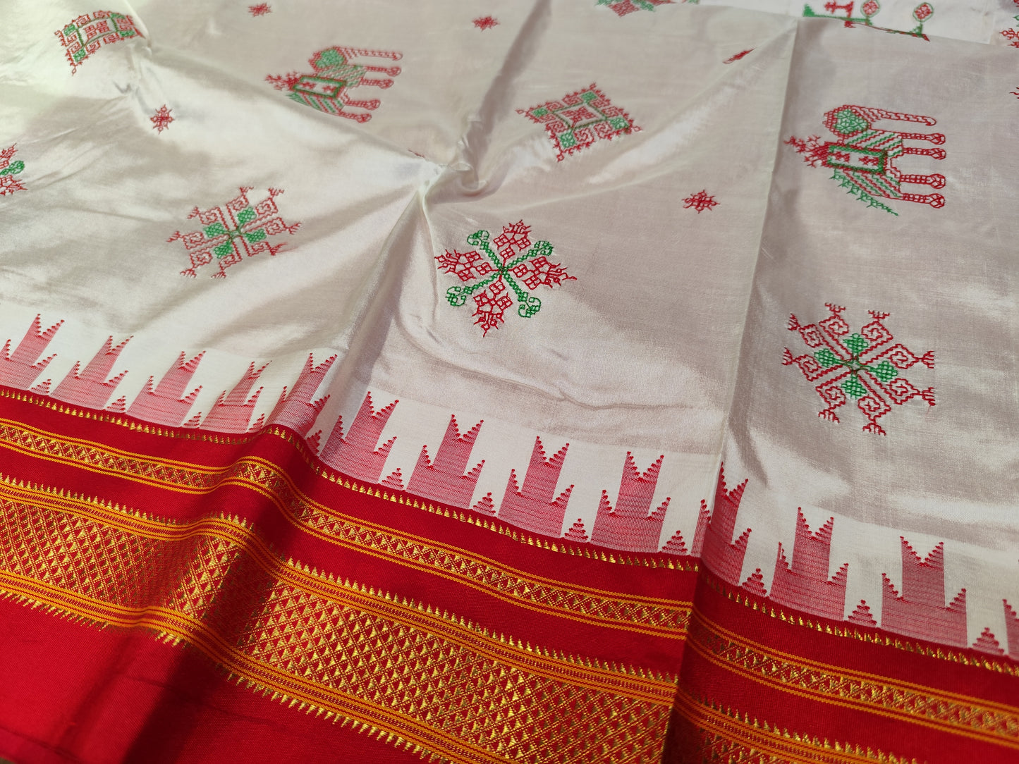 A close-up of a off white Ilkal silk saree with red and green embroidered motifs and a wide red border featuring gold zari-style detailing and a red geometric pattern.