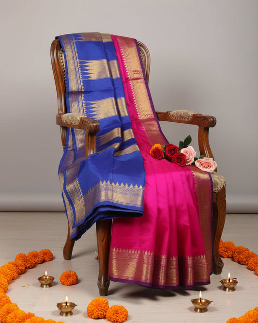 A vibrant blue and magenta pink silk saree with gold zari embroidery draped over a wooden armchair, styled with pink and red roses on the chair and floor.