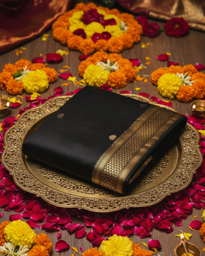 A folded black Kanjeevaram  silk saree with a wide gold and black woven border and small gold motifs, displayed on a light gray background.