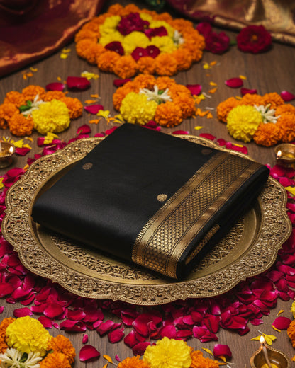 A folded black Kanjeevaram  silk saree with a wide gold and black woven border and small gold motifs, displayed on a light gray background.