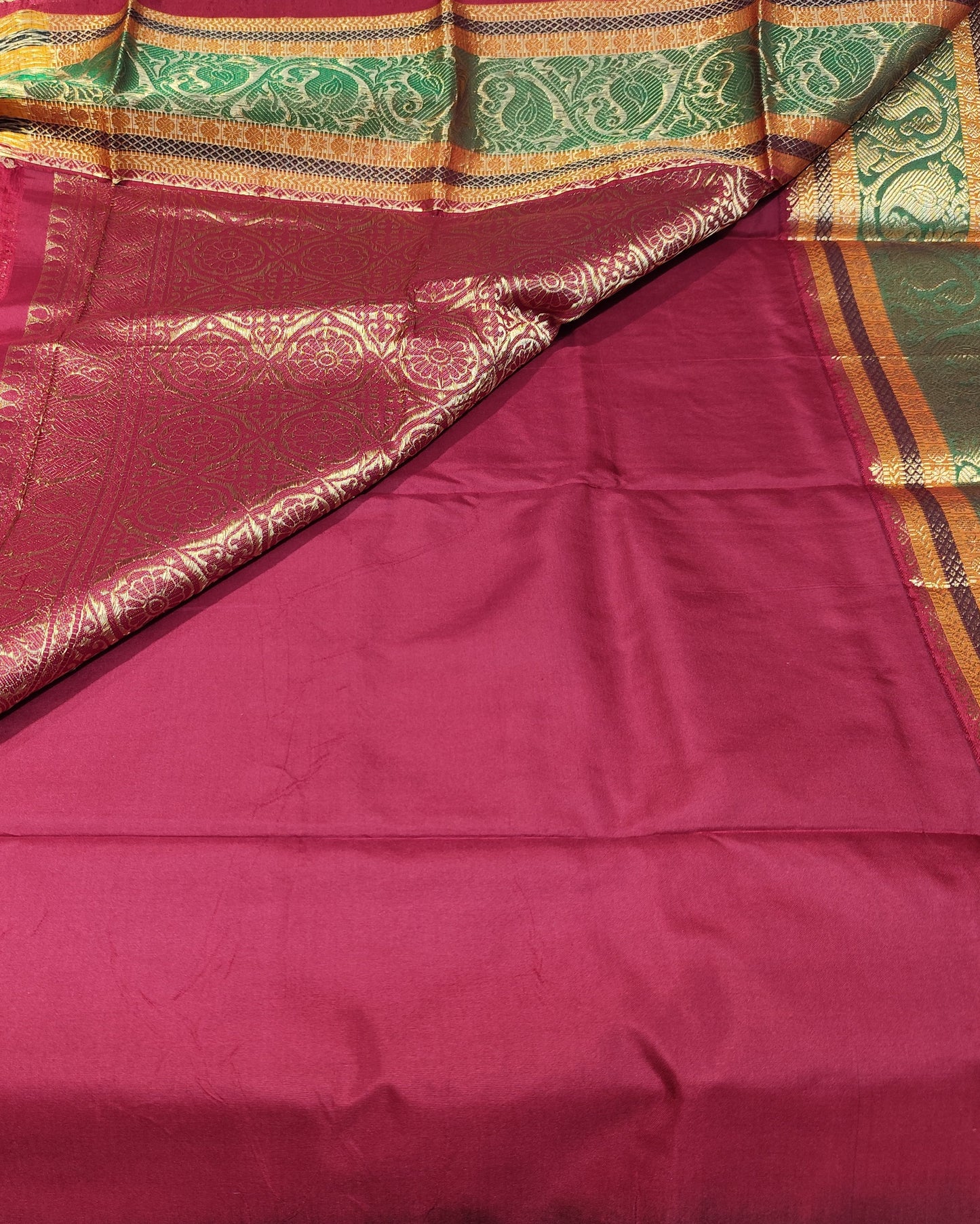 A deep red silk saree with intricate gold zari embroidery on the body and border, draped to show its patterned pallu and ornate green and gold decorative edges.