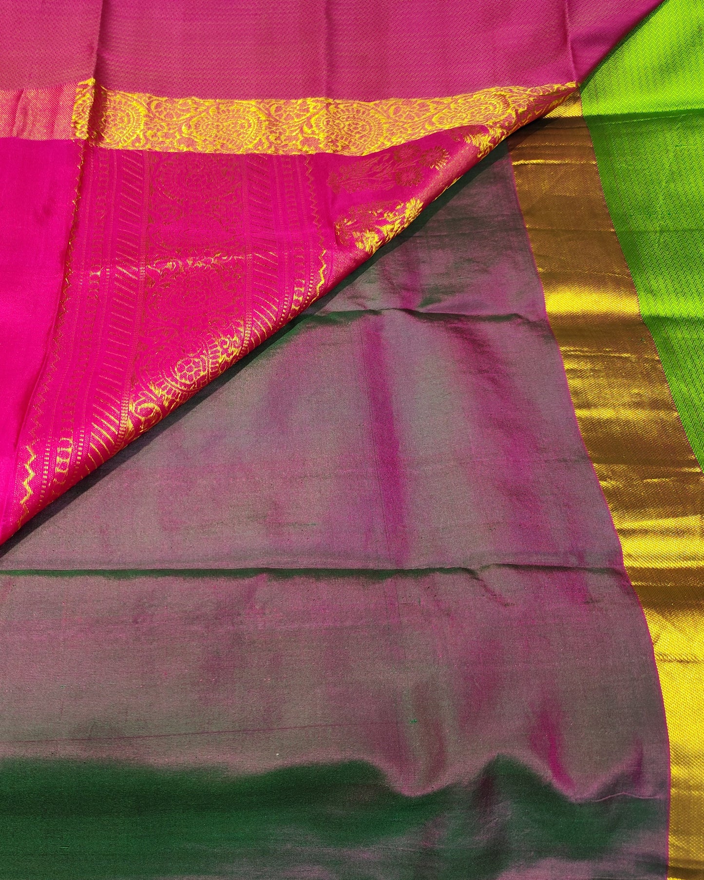 A folded and draped Kanjeevaram silk saree with a vibrant magenta-pink body, a dark green border, a bright lime green pallu, and ornate gold zari brocade patterns.