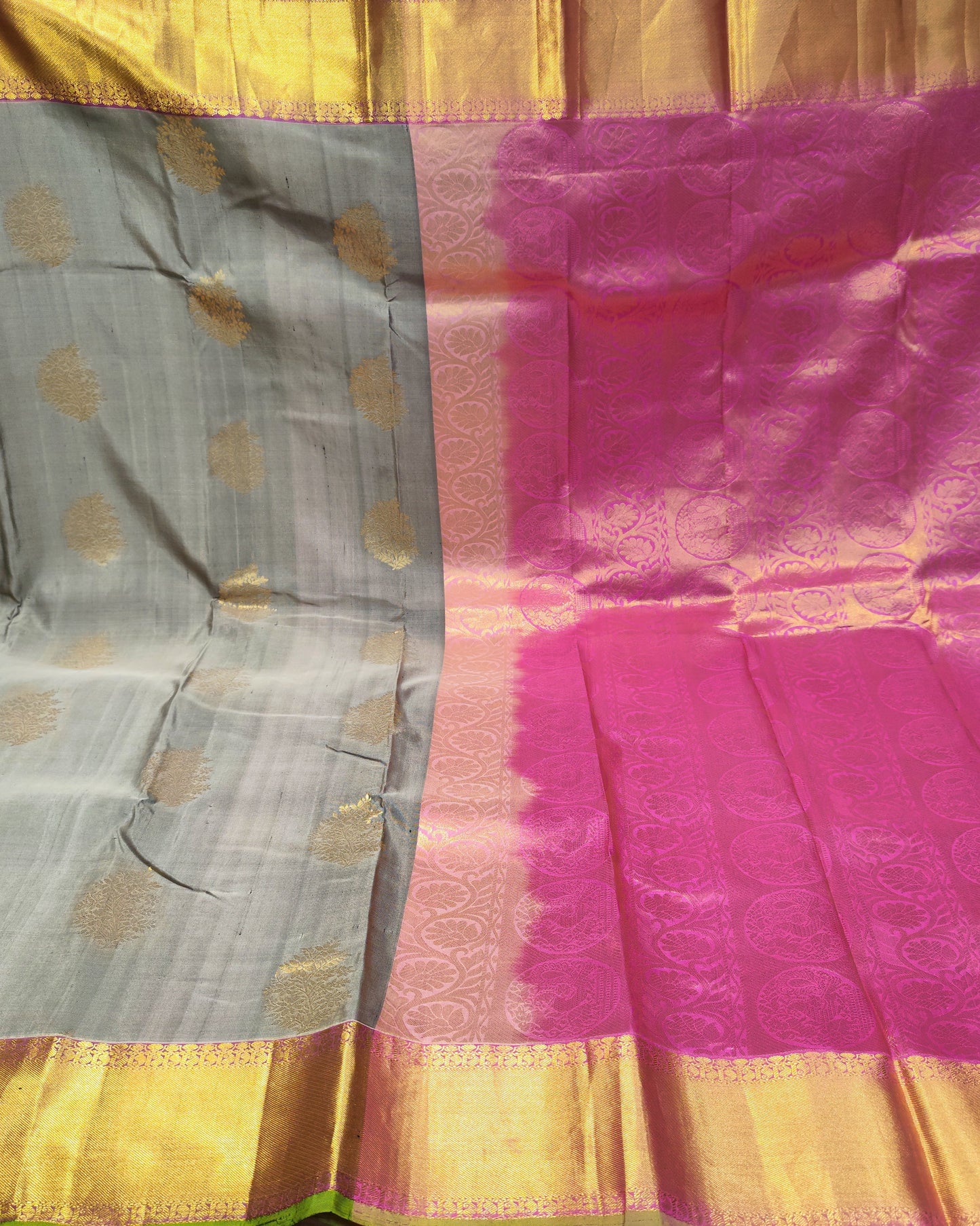 A folded and draped silk saree with a gray body, gold zari motifs, and a vibrant magenta-pink border with a wide gold zari pallu.