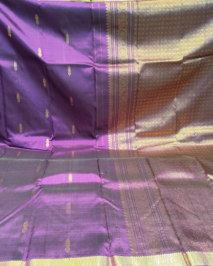 A purple silk Kanjeevaram saree with gold zari brocade work, displayed flat with the pallu section held up behind. The fabric is shown in a shop setting with other folded textiles visible in the background.