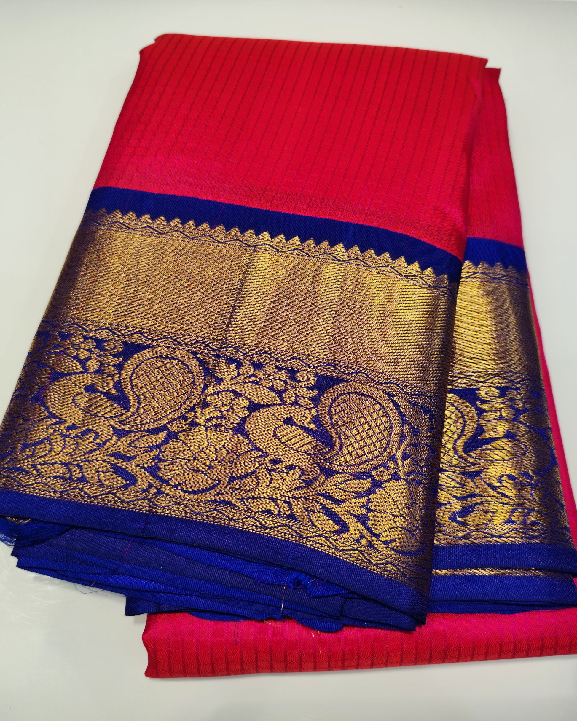 A folded red and blue saree with an ornate gold zari border featuring peacock motifs, displayed on a light gray surface.