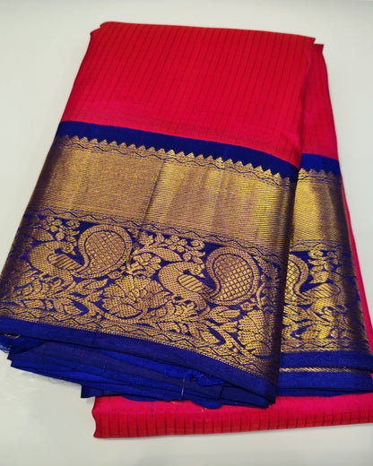 A folded red and blue saree with an ornate gold zari border featuring peacock motifs, displayed on a light gray surface.