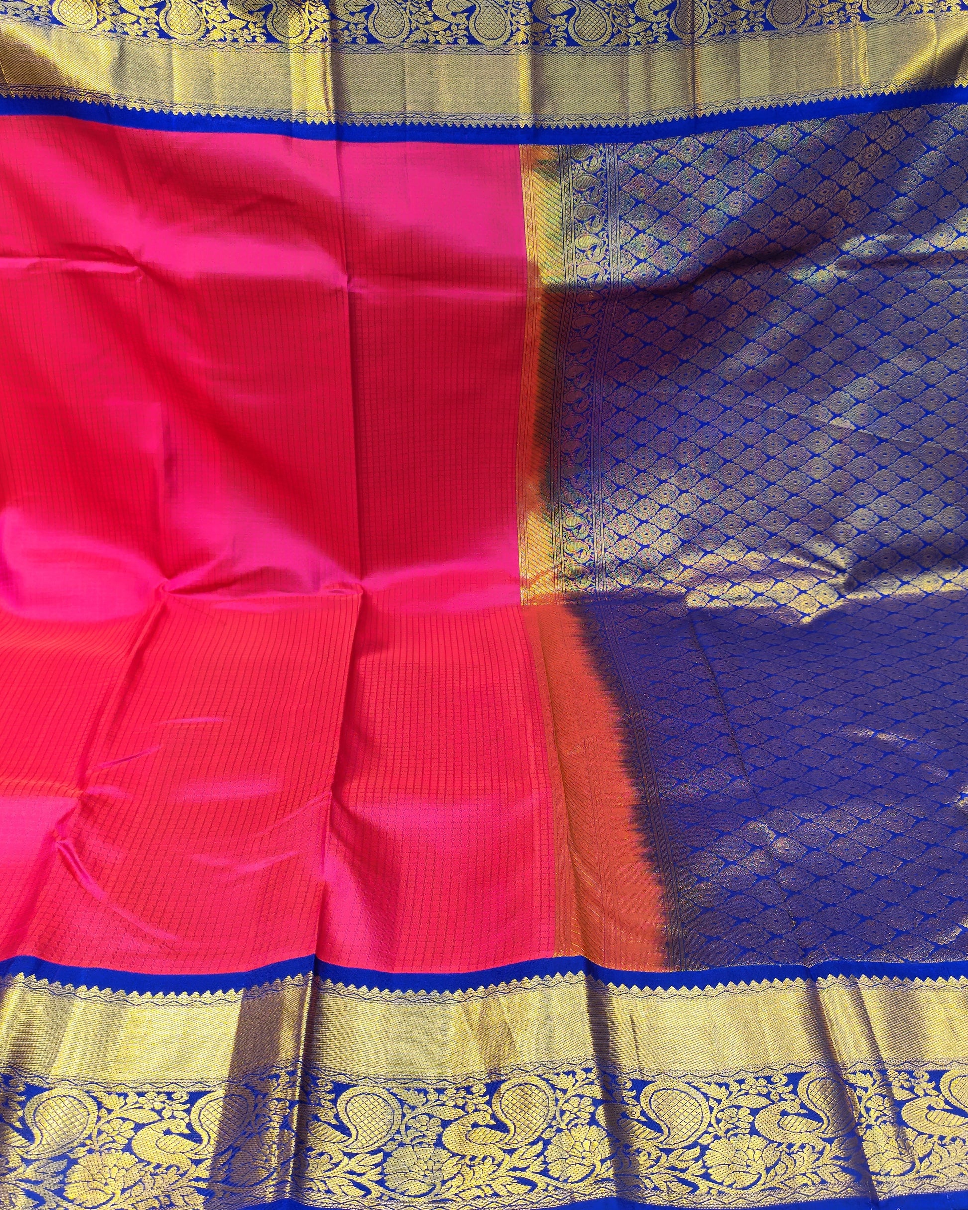 A vibrant pink and purple silk saree with intricate gold zari woven borders, displayed in a fabric store setting.