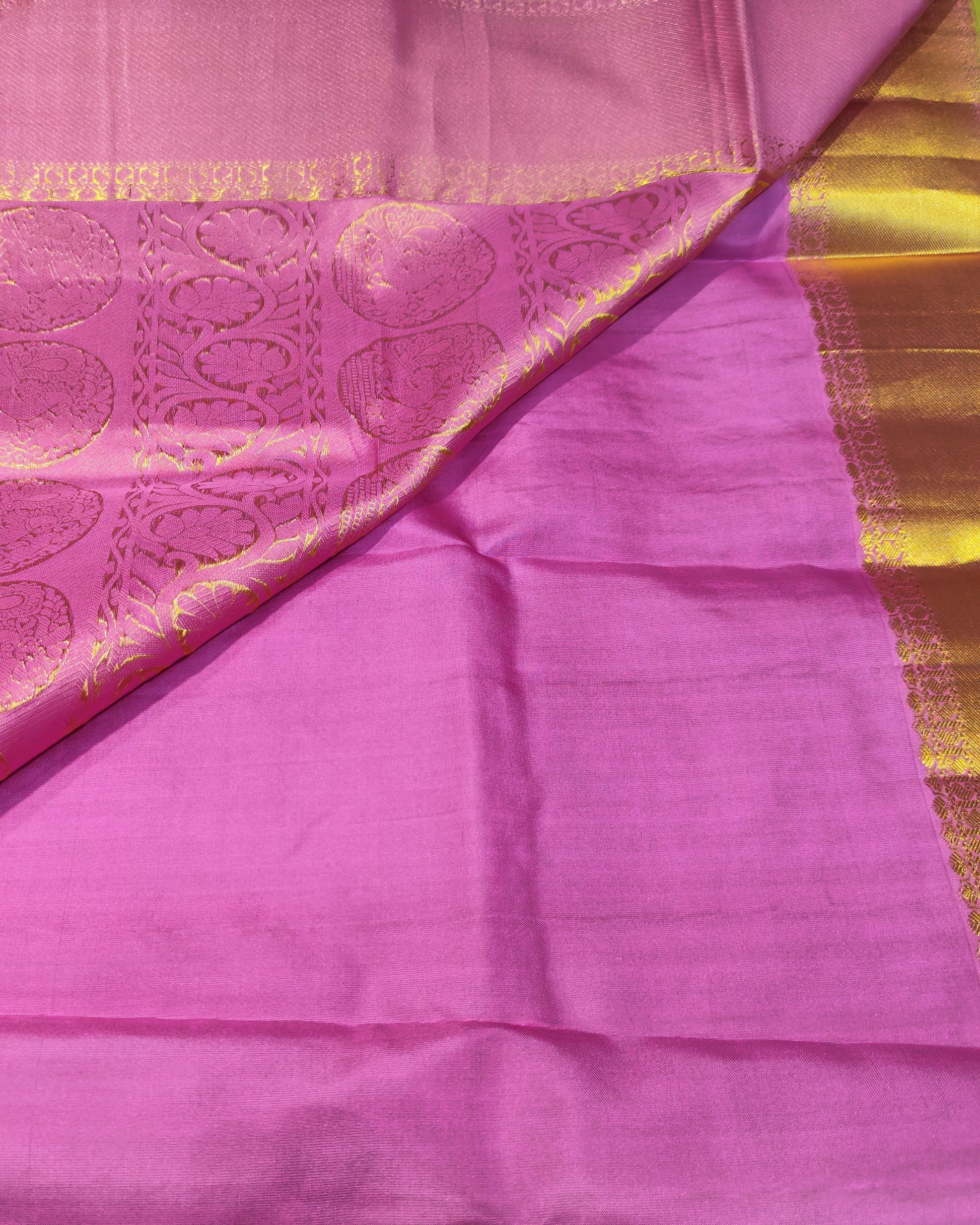 A pink silk saree with a wide gold zari border and matching gold patterned pallu, laid out on a white surface.