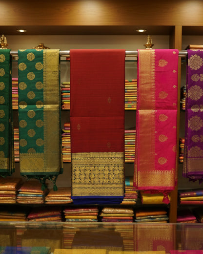A folded red silk saree with a wide gold and black zari border and a blue underlayer visible at the bottom, displayed on a light surface.
