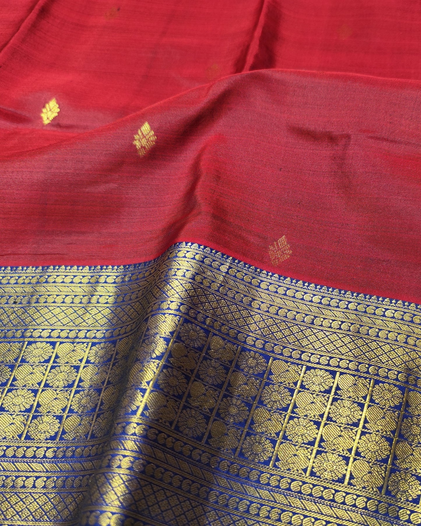 A close-up of a red silk saree fabric with a wide gold and blue zari border and small gold floral motifs woven on the red body.