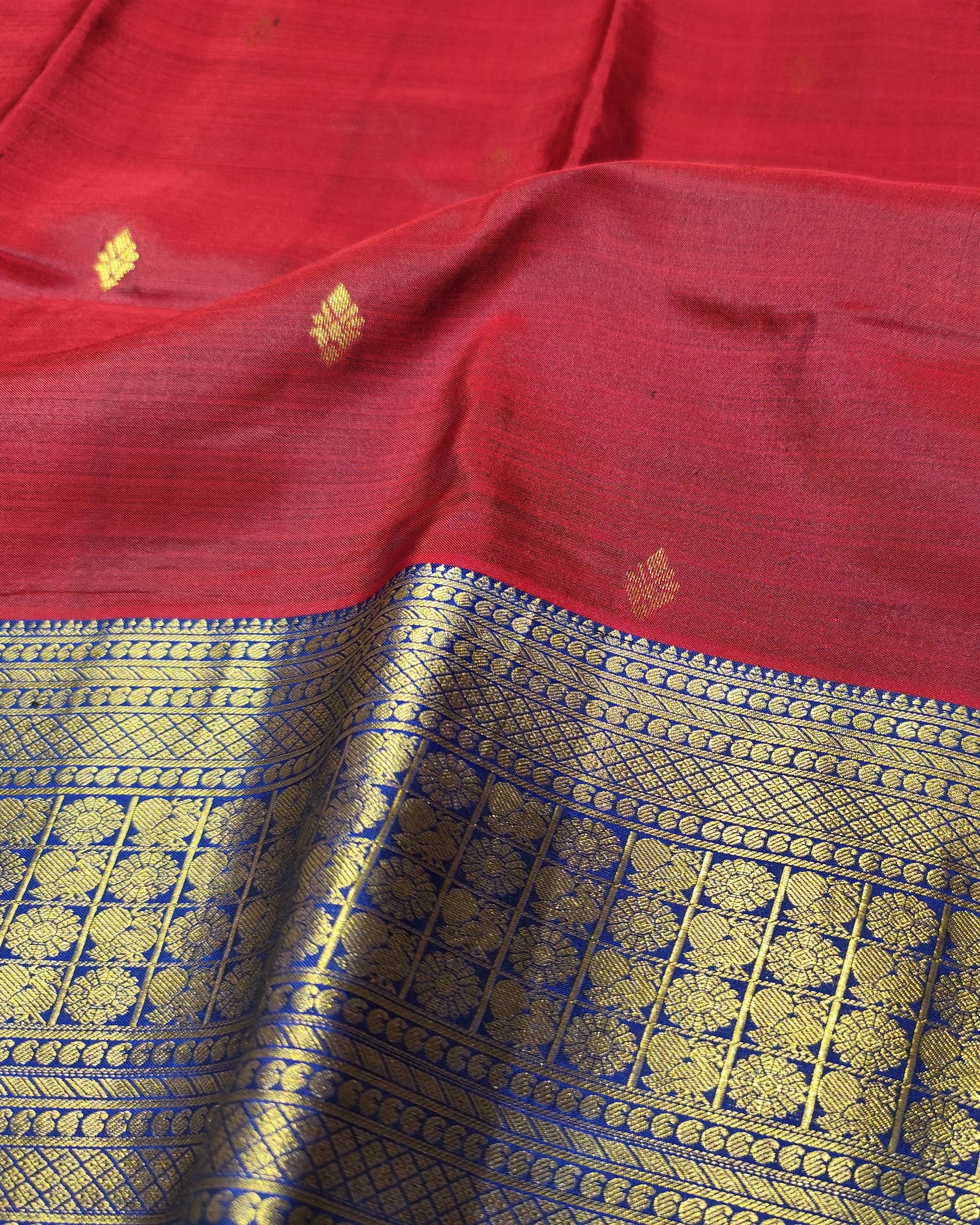 A close-up of a red silk saree fabric with a wide gold and blue zari border and small gold floral motifs woven on the red body.