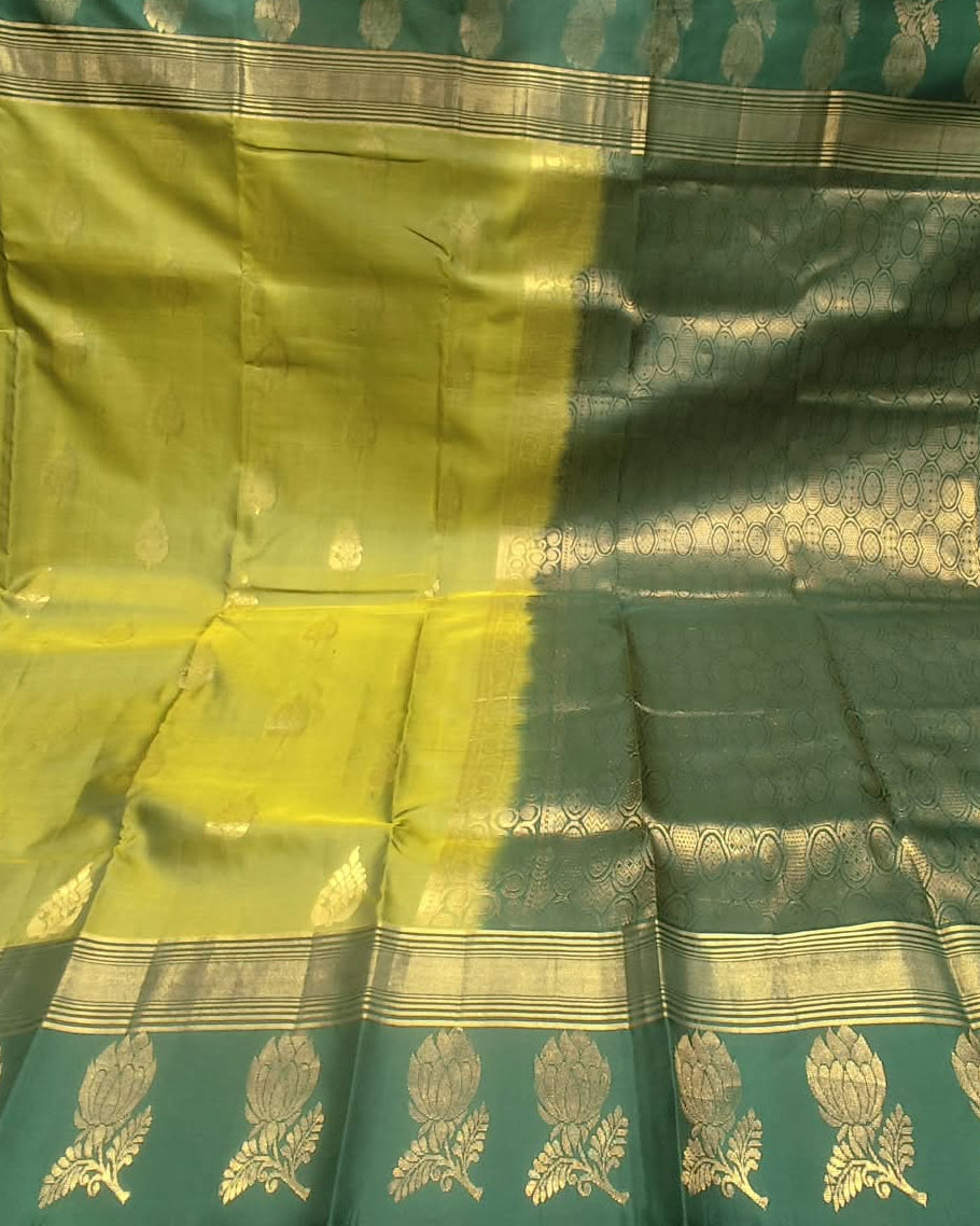 A green and yellow-green silk saree with intricate gold zari brocade patterns, displayed in a fabric store setting.