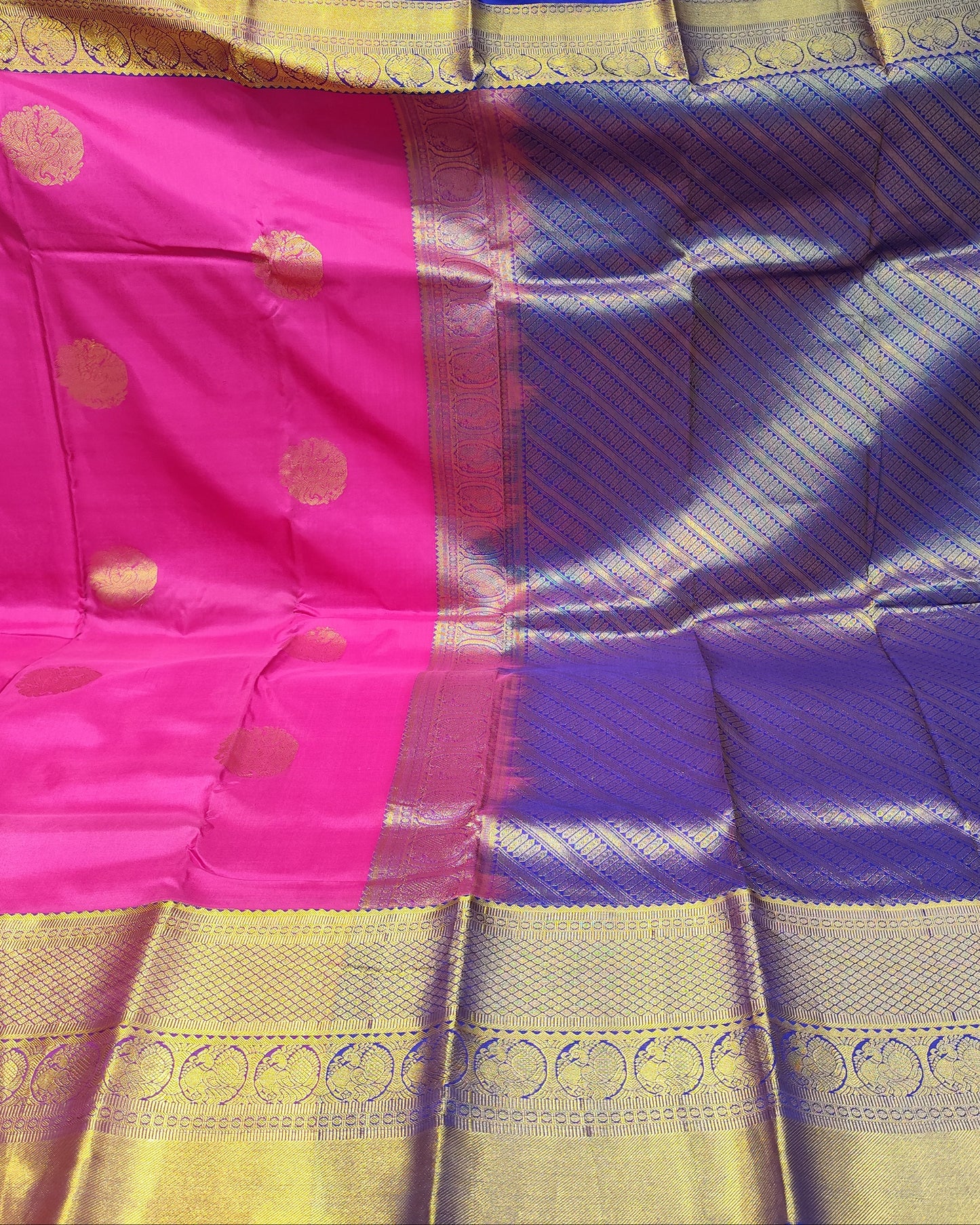 A folded and draped silk saree with a vibrant magenta pink body, a deep purple border, and a wide gold zari brocade border with metallic gold motifs.