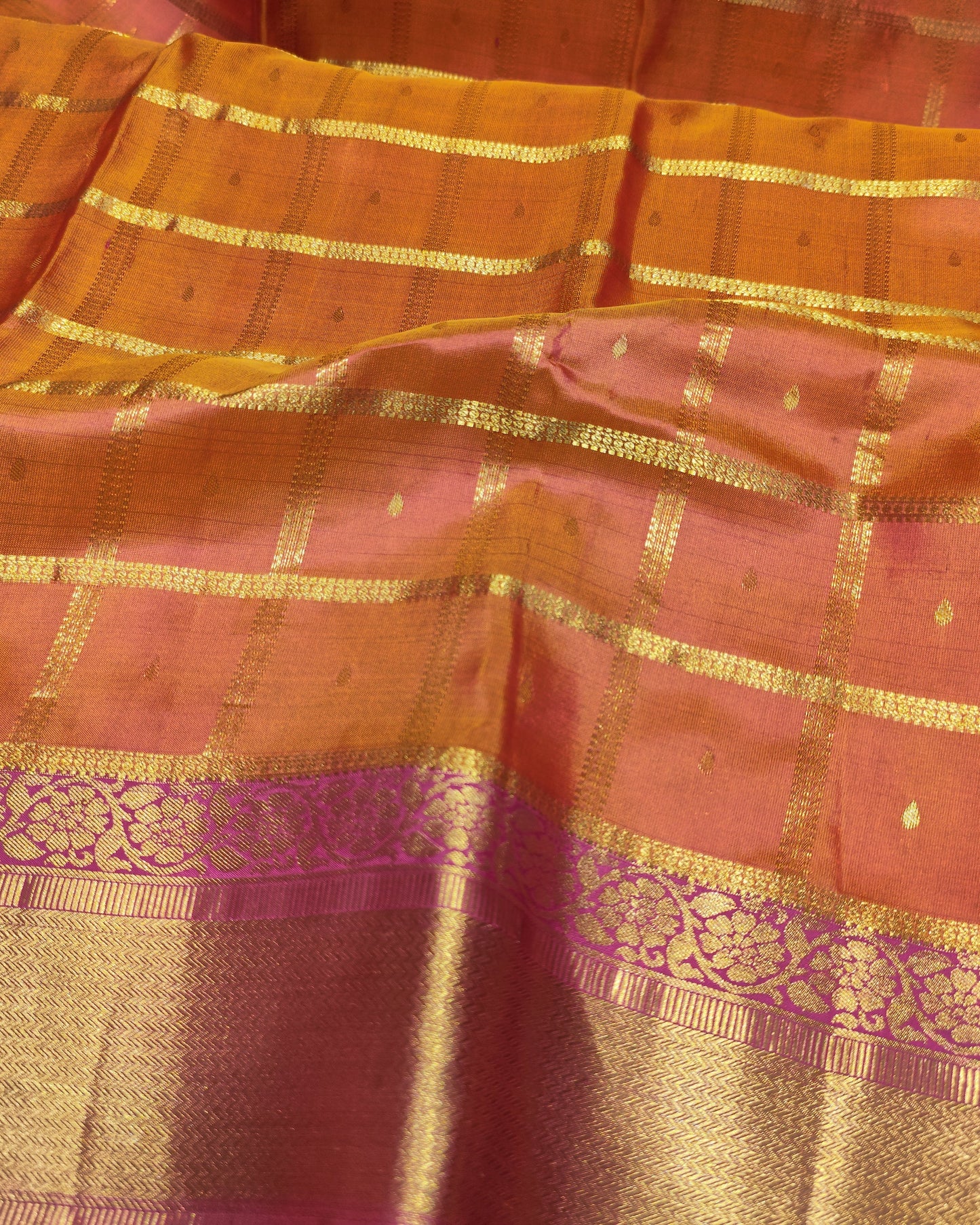 A close-up of a traditional Indian silk saree, showcasing its vibrant orange and pink body with intricate gold zari woven checkered patterns and a wide decorative gold border.