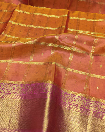 A close-up of a traditional Indian silk saree, showcasing its vibrant orange and pink body with intricate gold zari woven checkered patterns and a wide decorative gold border.