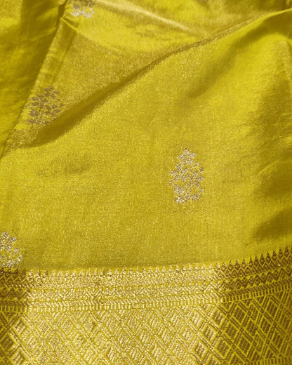A close-up of a bright yellow-green silk fabric, likely a saree, featuring a wide gold zari brocade border and small metallic floral motifs woven throughout.