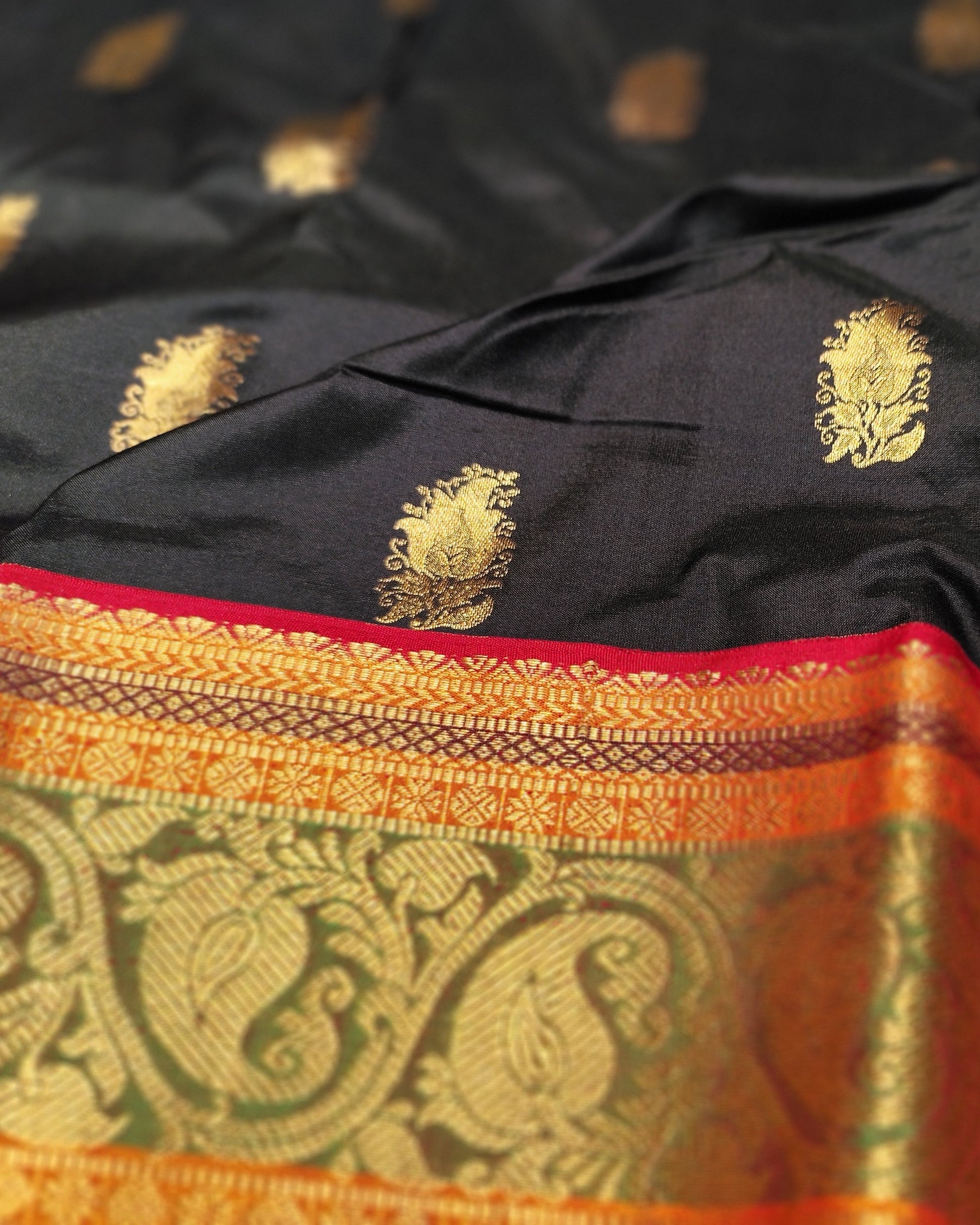 A close-up of a black silk saree with gold zari motifs and a wide orange and gold brocade border with red piping.
