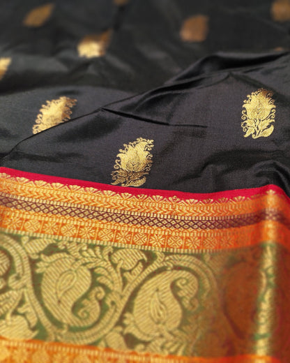 A close-up of a black silk saree with gold zari motifs and a wide orange and gold brocade border with red piping.